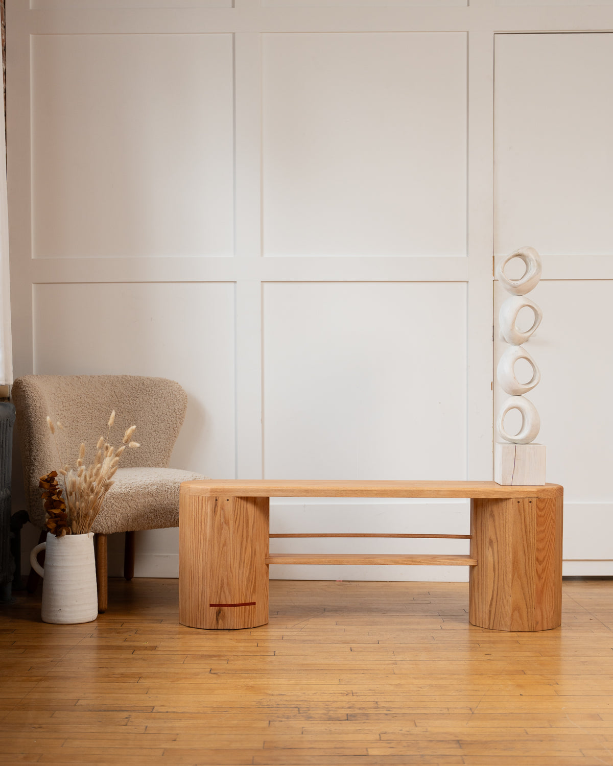 Handmade red oak iB-v2 Dual Column Bench by Redline Design with its signature red line, beside a boucle chair